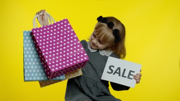 Menina criança alegre bonito na camisa mostrando venda palavra anúncio inscrição banner e sacos de compras - Filmagem, Vídeo