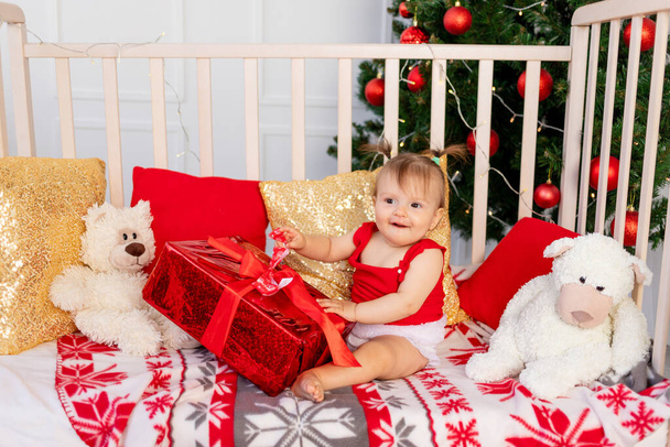 bebê pequeno feliz seis meses em um berço em casa pela árvore de Natal com um grande presente vermelho, ano novo e conceito de feriado - Foto, Imagem