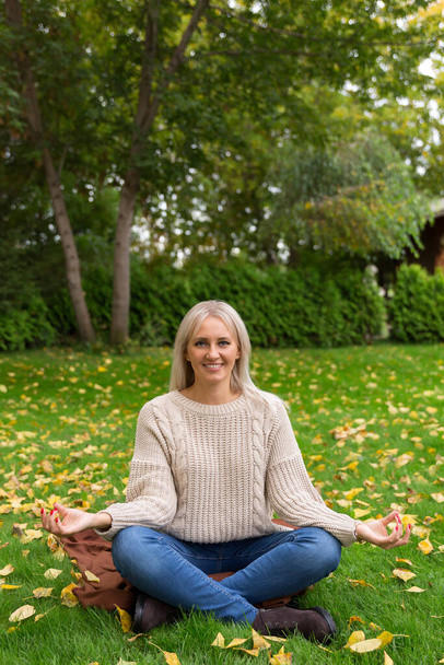 lawn is covered with yellow autumn leaves. Woman sitting cross legged in lotus position and meditating, on green grass. - Photo, image