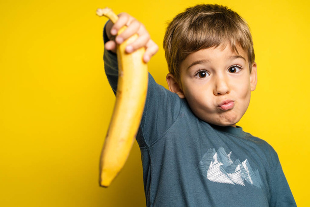 Portrait of happy small caucasian boy in front of yellow background holding banana making face - Childhood growing up and food concept - front view waist up copy space - Fotó, kép