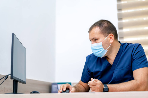 Νεαρός όμορφος άντρας γιατρός διαβάζει ιατρικά αποτελέσματα στην οθόνη. Σύγχρονο νοσοκομειακό γραφείο. - Φωτογραφία, εικόνα