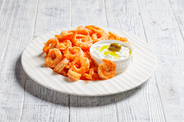 Seafood plate of fried garlic prawns or shrimps deshelled and deveined served with greek tzatziki sauce, green olives, rosemary on a white plate on a white wooden background, top view, close-up - Zdjęcie, obraz