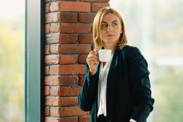 Business woman holding a cup of coffee looking to the side. Young elegant blonde standing by the window in the office. Enjoy coffee during your work break. High quality photo - Fotoğraf, Görsel