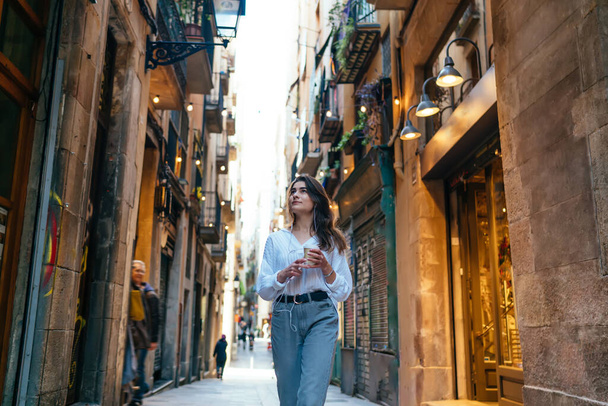 Продумана молода жінка в повсякденному одязі, що ходить по старій вулиці, п'є каву і слухає музику на смартфоні
 - Фото, зображення