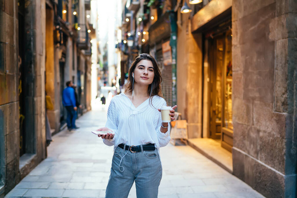 Thoughtful hipster girl in electronic earphones looking away while waiting guide for travel sightseeing, Caucasian female tourist with coffee to go and cellular technology listening online book - Foto, Imagem