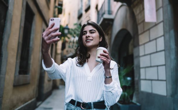 Pretty Caucasian woman clicking selfie pictures during travel sightseeing using front mobility camera, happy hipster girl with coffee to go smiling while shooting influence vlog video about tourism - Fotó, kép