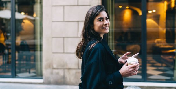 Side view of cheerful dark haired woman in black coat holding cup of coffee to go and smartphone and looking at camera - Photo, Image