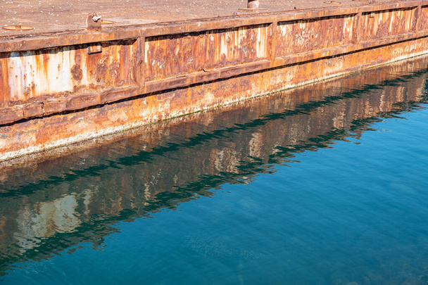 澄んだ青い海の水に錆びた金属製の古い工業港の岸壁反射. - 写真・画像