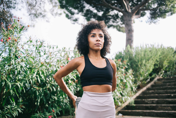 Confident ethnic sporty female with curly hair wearing sportswear stretching legs and looking away while standing in green garden near stairs - Foto, Imagem