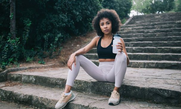 Tired young slim ethnic sportswoman in activewear and sneakers relaxing on stairs of park with bottle of water during workout - Zdjęcie, obraz