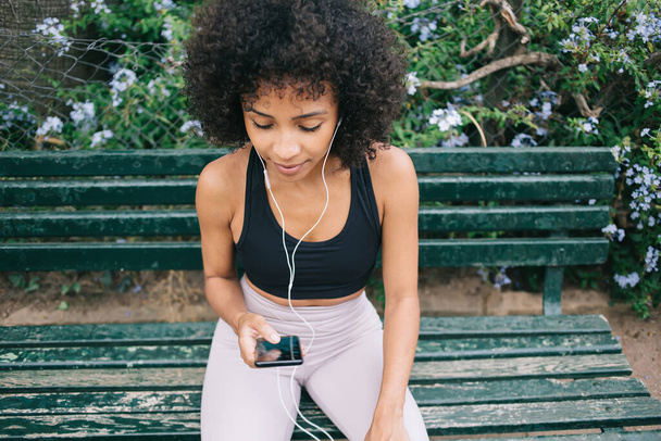 From above of young sporty ethnic female in activewear with earphones and smartphone listening to songs while sitting on bench in park - Фото, изображение