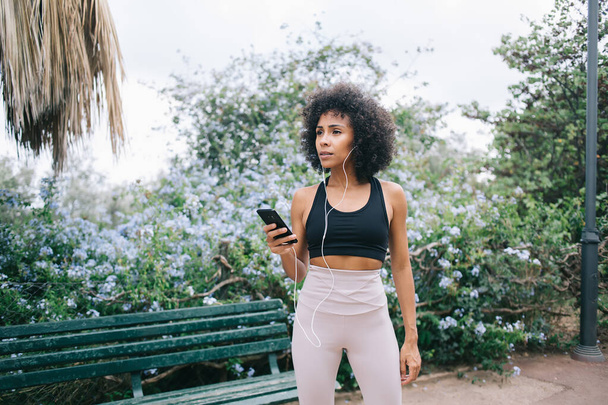 Focused ethnic young sportswoman with curly hair in activewear listening to music and browsing smartphone while standing near bench in floral park - Photo, image