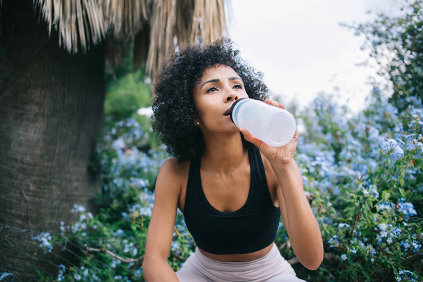 Thirsty African American lady with Afro hairstyle enjoying fresh water while having break in workout in park near blossoming bushes - Foto, Bild