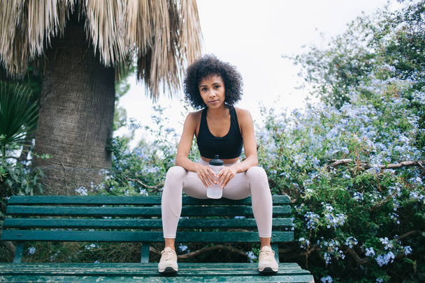 Volle lengte zelfverzekerde etnische vrouw in sportkleding met Afro kapsel leunend op knieën op bank in stadspark onder palm in de buurt van bloeiende bomen met plastic waterfles - Foto, afbeelding