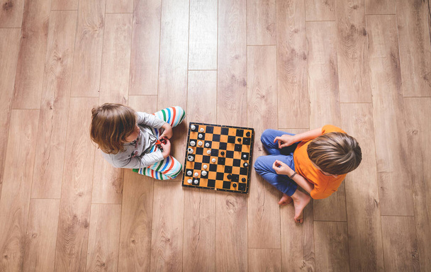 床の上でチェスをしている子供の男の子と女の子は激しく考えています。テーブルゲームをしている子供たち。家庭での教育。学校概念. - 写真・画像
