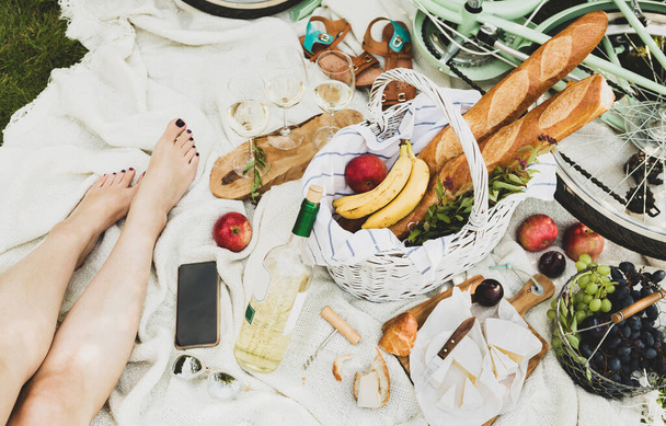French style outdoor picnic on the white blanket. Barefoot woman, wicker basket with baguettes, wine, fruits and cheese around. Pastel green bicycle in the background. Summer leisure - lifestyle. - Foto, Bild