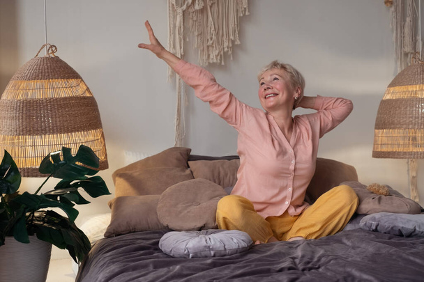 Kaukasische Seniorin im Pyjama lächelt im hellen Wohnzimmer und streckt sich beim Aufwachen. - Foto, Bild