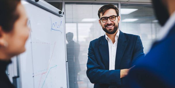 Cheerful male leader of company smiling near flip chart during presentation with colleagues, happy businessman in formal outfit and spectacles discussing economic graphics with cropped partners - Foto, Imagen