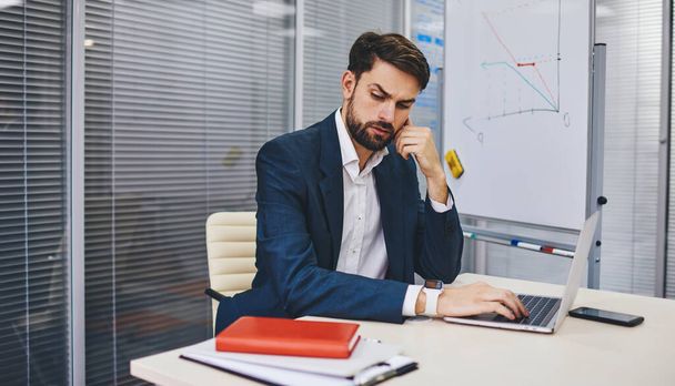 Pensive male director dressed in formal suit thoughtful looking away while sitting at desktop with laptop computer and creating business project, executive manager puzzled on web info in office - Photo, Image