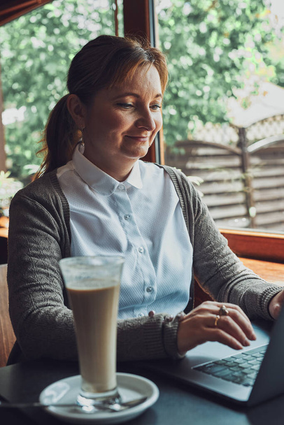 Улыбающаяся счастливая деловая женщина, работающая удаленно на ноутбуке, управляющая своей работой, печатая на клавиатуре ноутбука, сидя за столом в кафе - Фото, изображение