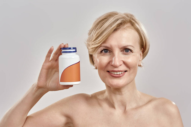 Portrait of middle aged caucasian woman holding a bottle with pills, smiling at camera and standing isolated over grey background. Healthcare concept - Fotoğraf, Görsel