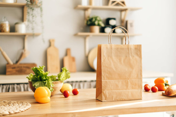 Eco shopping paper bag with fresh vegetables and baguette on the table in modern kitchen. Food delivery or market shopping concept. - Fotó, kép