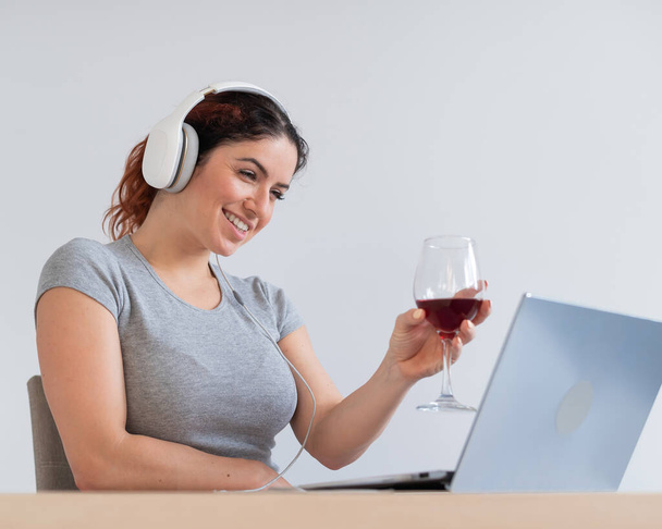 Μια γυναίκα με ακουστικά πίνει κόκκινο κρασί και επικοινωνεί με φίλους μέσω βίντεο επικοινωνίας σε ένα φορητό υπολογιστή. Online chat σε καραντίνα - Φωτογραφία, εικόνα