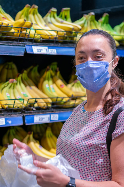 Mujer enfocada en mascarilla facial eligiendo frutas, tomando plátanos de los estantes en la tienda de comestibles. Cliente en el supermercado. Vista lateral. Compras durante el concepto epidémico. foto vertical - Foto, Imagen