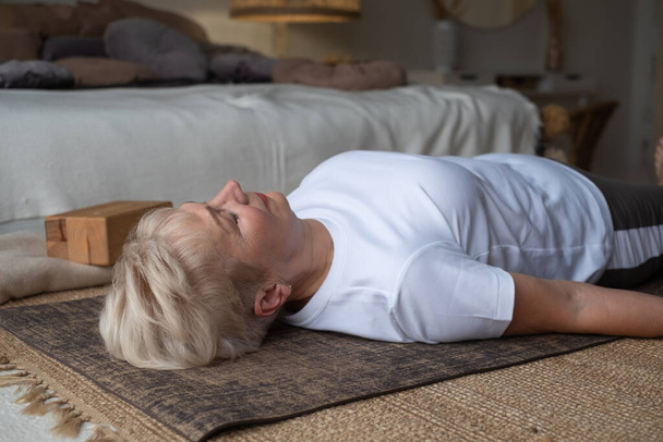Vanhempi valkoihoinen nainen makaa joogamatolla treenin jälkeen. Sopii naaras rentouttava lattialla kotona. - Valokuva, kuva