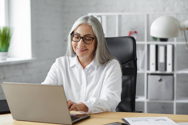 Grey-haired senior businessman working in office with laptop. Business portrait of beautiful smiling elderly woman wearing eyeglasses sitting at workplace. Mature people with computers - 写真・画像