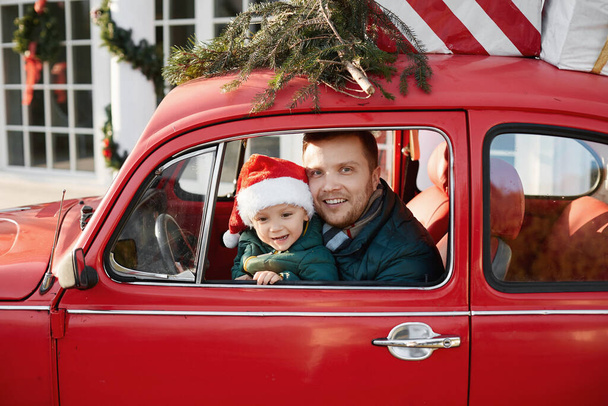 Handsome man with cute baby boy in Santa Claus hat inside the red retro car with Christmas gifts - Fotó, kép