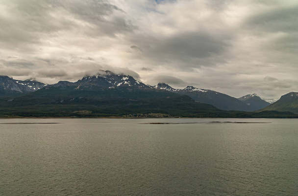 Tierra del Fuego, Argentina - December 13, 2008: West side Beagle Channel. Line of small islets in dark gray water in front of snowy mountains under thick gray clooudscape with lighter spots. - Photo, Image