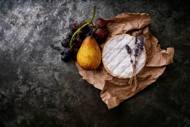 Ranskalainen juusto Camembert hedelmiä ja kuivattua laventelia varret paperilla tumman maalaismainen tausta. Huippunäkymä. Kopioi tila - Valokuva, kuva