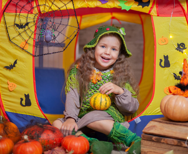 A little cute girl dressed as a forest fairy sits in a children's tent decorated for Halloween with bats and ghosts, a spider web with a spider, orange pumpkins. - 写真・画像