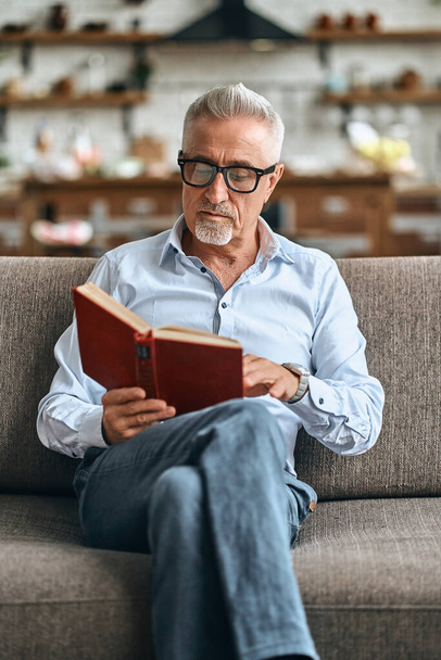 Elegante homem maduro ler livro depois de trabalhar duro dia enquanto sentado no sofá acolhedor. Conceito de estilo de vida doméstico - Foto, Imagem