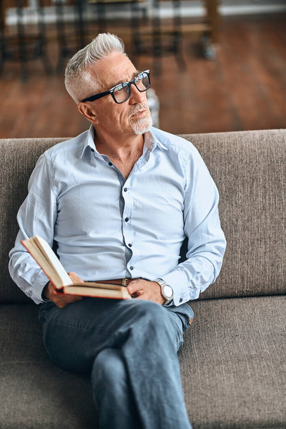 Gray-haired mature man in a blue shirt resting on the sofa and holding a book in the living room. Domestic lifestyle concept - Fotografie, Obrázek