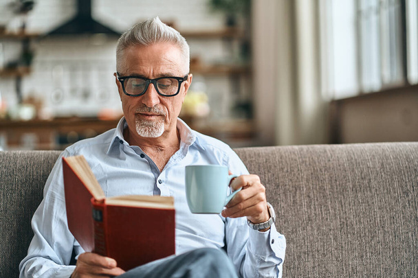 Vyötärö ylös kypsä mies lukee kirjaa istuessaan sohvalla olohuoneessaan ja pitämällä kuppi kahvia. Kotimaisen elämäntavan käsite. Kopioi tila - Valokuva, kuva