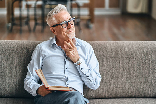 Smiling mature male looking away and dreaming with a book in his hand in living room at home. Domestic lifestyle concept. Copy space - 写真・画像