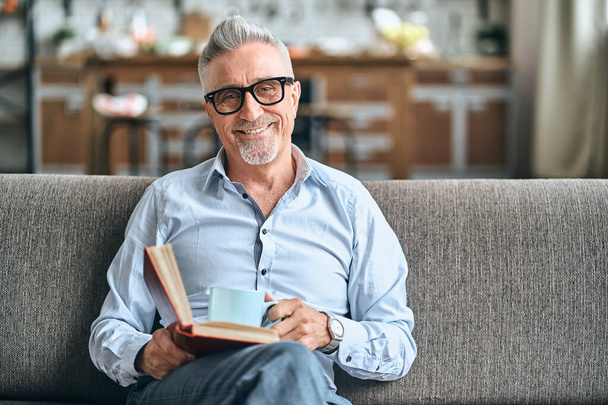 Smiling bearded mature man in a blue shirt resting on sofa while holding book and cup of hot drink at home. Domestic lifestyle concept. Copy space - 写真・画像