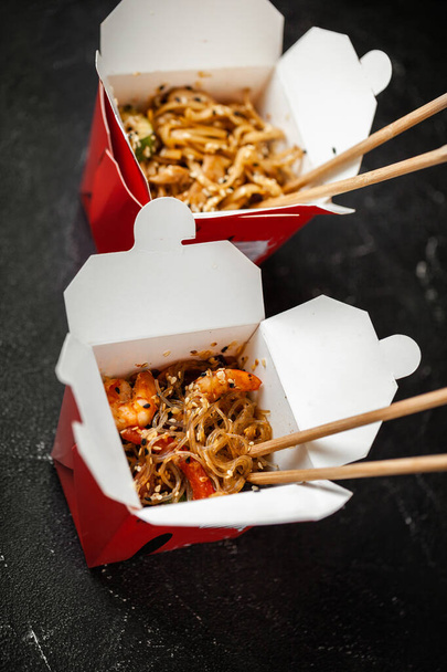 Udon noodles with fried chicken and vegetables in the cardboard box with wooden chopsticks on the black vintage background - Photo, image
