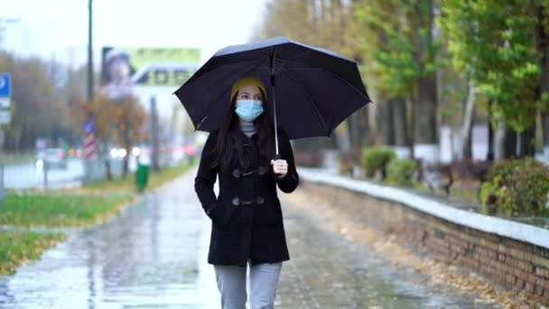 Μια νεαρή γυναίκα με προστατευτική μάσκα που περπατάει στο πάρκο κάτω από ομπρέλα. Βροχερή μέρα, κατά τη διάρκεια της δεύτερης καραντίνας κύμα coronavirus COVID-19 πανδημία - Πλάνα, βίντεο