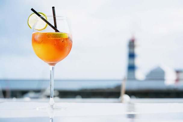 Aperol spritz, Italian summer cocktail against the lighthouse and seascape background, outdoor cocktail bar. Selective focus - Photo, Image