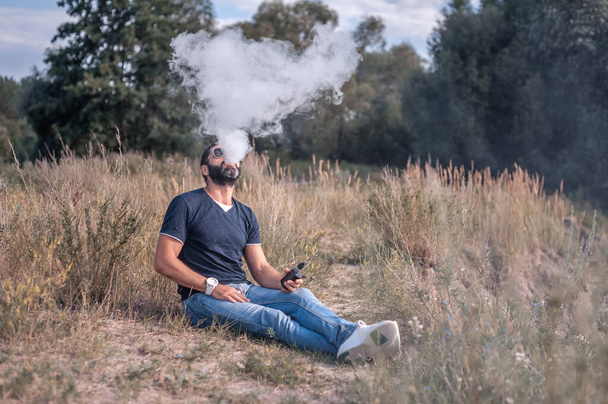Hombre de vapor guapo disfrutando de un dispositivo de humo electrónico en la hierba. Fumar no tabaco. - Foto, imagen