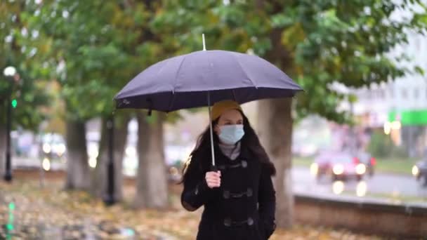 Parkta şemsiye altında yürüyen koruyucu maskeli genç bir kadın. Yağmurlu bir günde, ikinci dalga karantina sırasında koronavirüs COVID-19 salgını - Video, Çekim