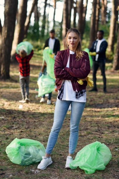 Active pretty girl with full rubbish bags in backdrop of her friends volunteers which are picking up rubbish at forest. - Foto, Imagen