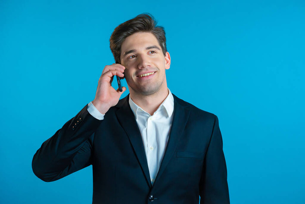 Young businessman speaks with smile on phone. Guy holding and using smart phone. Suit outfit. Blue studio background. - Fotó, kép