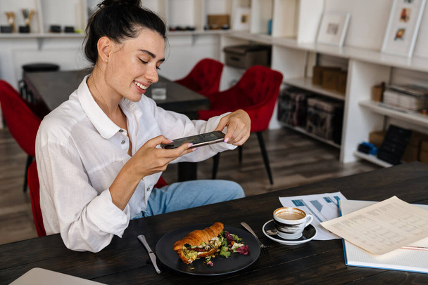 Attractive happy young woman taking photo of delicious breakfast with croissants with smartphone in cafe - Photo, image