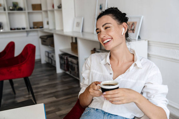 Happy young woman relaxing in cafe, wearing wireless earphones, sitting at the tablewith cup of coffee - Foto, Imagem