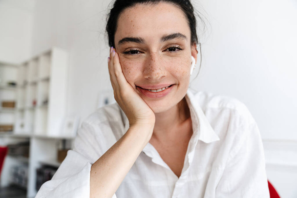 Close up of a happy young woman on a video call, looking a camera, wearing wireless earphones - Photo, Image