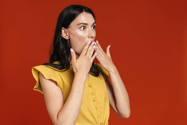 Изображение молодой шокированной женщины, выражающей удивление и закрывающей рот на красном фоне - Фото, изображение